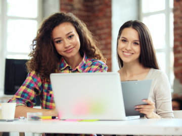 Zwei junge Frauen sitzen gemeinsam an einem Laptop und blicken zum Betrachter.