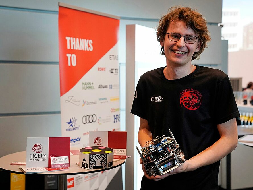 Lachender Student mit Fußballroboter in einer Größe von ca. 20 cm Höhe in der Hand.