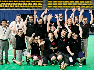 Gruppenfoto mit 13 Studierenden der DHBW Mannheim, die bei der Award Ceremony auf einem Roboterfußballfeld jubelnd die Arme hochreißen. 