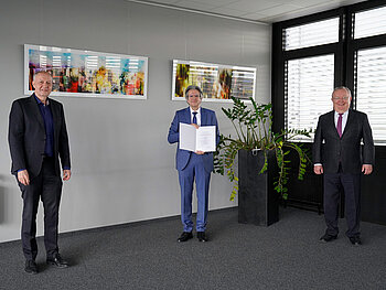 3 Männer im Anzug in einem Konferenzsaal mit der Urkunde zum Dienstjubiläum