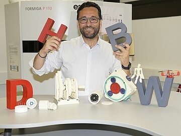 Porträt Prof. Dr. Hulusi Bozkurt im Labor für Additive Fertigung mit 3D-gedruckten Kunststoffteilen und den Buchstaben DHBW in der Hand.