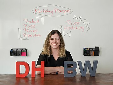 Porträt Helena Martin vor einem Whiteboard mit Begriffen zu ihrer Doktorarbeit; sie lehnt an einer Theke mit den 4 DHBW-Buchstaben.