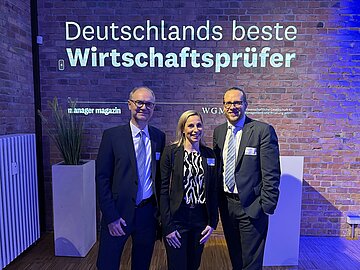 Gruppenbild mit drei Personen, im Hintergrund an der Wand die Projektion "Deutschlands beste Wirtschaftsprüfer" in blau und lila Licht getaucht. 