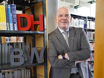 Porträt Michael Lang lehnend an einem Bücherregal in der Bibliothek der DHBW Mannheim, im Regal stehen die Buchstaben DHBW.