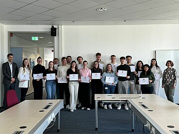 Gruppenbild mit 18 Studierenden, die ihr Zertifikat halten in einem Schulungsraum. 