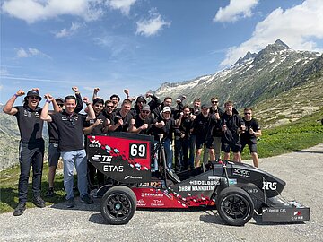 Gruppenbild mit jubelnden Personen und Rennwagen, im Hintergrund Schweizer Berge und eine Wiese. 