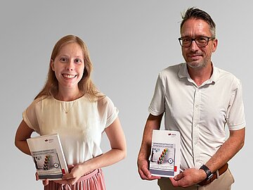 Studentin links, rechts daneben der Professor, beide sommerlich gekleidet und halten jeweils das Buch in der Hand. 