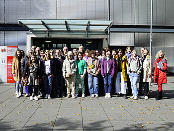 Besuch der Berater*innen der Agenturen für Arbeit aus der Metropolregion Rhein-Neckar und aus Darmstadt am 23.09.2025.  Gruppenbild im herbstlichen Sonnenschein mit 40 Personen auf dem Außengelände der DHBW Mannheim; hinter den in Jacken gehüllten Personen der Eingang zu Gebäude D mit heruntergefahrenen Jalousien.