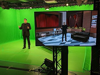 Prof. Dr. Dinkel führt das Greenscreen Innovation & Applications Lab vor Prof. Dr. Dinkel steht im Greenscreen-Labor, rechts voran steht ein Bildschirm, auf dem er vor einem virtuellen Hintergrund gezeigt wird.
