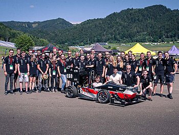 CURE-Mitglieder stehen mit Rennauto vor einem Bergpanorama