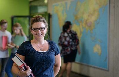 Studentin an der DHBW Mannheim mit Büchern in der Hand schaut in die Kamera, im Hintergrund sieht man Studierende vor einer Weltkarte