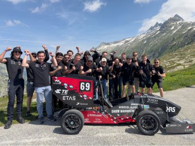 17 jubelnde Studierende hinter ihrem elektrischen Rennwagen, im Hintergrund Weiden und Berge und sonniger Himmel