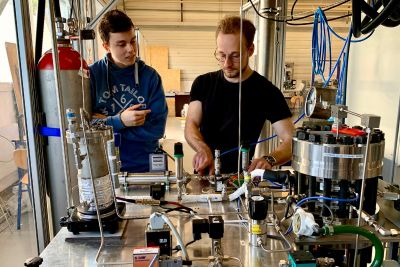 DHBW-Studierende arbeiten im Wasserstofflabor an einem elektrochemischen Verdichter. Damit lässt sich H2 in einem Schritt reinigen, verdichten und in Druckflaschen bei 200 bar speichern. Zwei Studierende mit einem Elektrochemischen Verdichter im Wasserstoff-Labor der DHBW Mannheim, neben ihnen eine Wasserstoff-Flasche.
