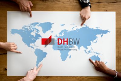 Weltkarte auf einem großen weißen Blatt Papier, rundherum 5 Hände, deren Zeigefinger auf unterschiedliche Orte der Welt zeigen. In der Mitte der Karte das DHBW-Logo. 